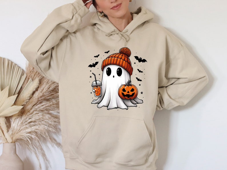 Pu&ograve; includere: Felpa con cappuccio beige con un fantasma dei cartoni animati con un berretto arancione, che tiene in mano una zucca e una bevanda. Pipistrelli e stelle nere circondano il fantasma. Perfetto per Halloween.