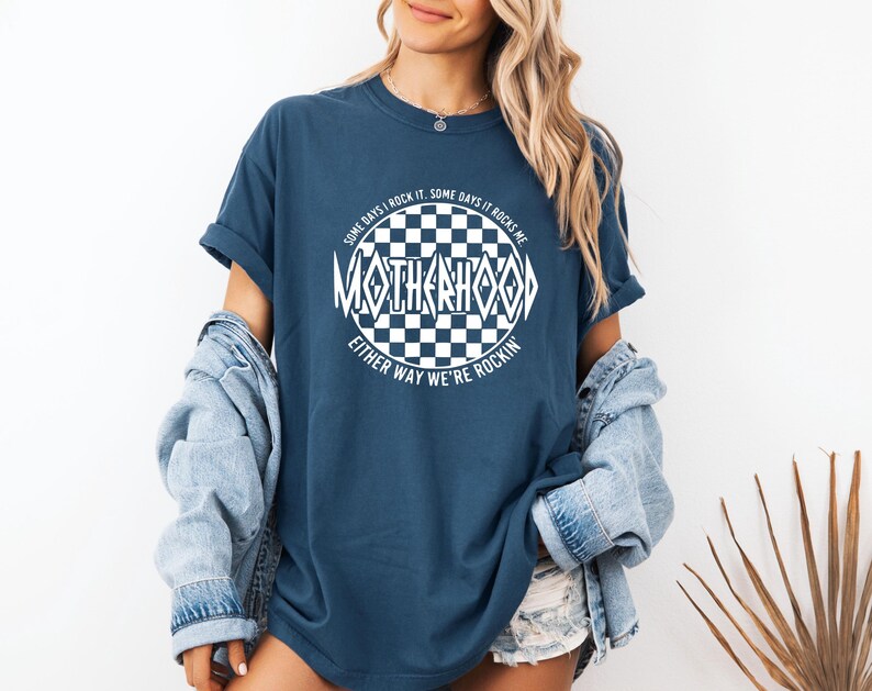 May include: A navy blue t-shirt with a white checkered circle graphic that says "Some days I rock it. Some days it rocks me. Either way we're rockin'." The text inside the circle says "Motherhood".