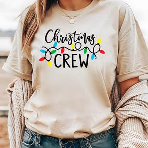 Puede incluir: Camiseta beige con la frase "Christmas CREW" en negro, adornada con una guirnalda de luces navide&ntilde;as de colores. La camiseta tiene mangas cortas y un corte relajado, ideal para la temporada navide&ntilde;a.
