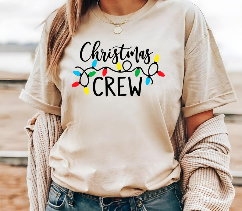 Puede incluir: Camiseta beige con la frase "Christmas CREW" en negro, adornada con una guirnalda de luces navide&ntilde;as de colores. La camiseta tiene mangas cortas y un corte relajado, ideal para la temporada navide&ntilde;a.