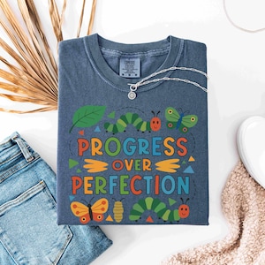 May include: A blue t-shirt with the words "PROGRESS OVER PERFECTION" in colorful letters and a whimsical design of caterpillars, butterflies, and leaves. A silver necklace and white shoes are also visible.