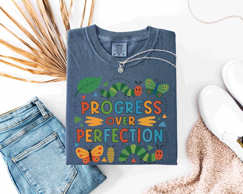 May include: A blue t-shirt with the words "PROGRESS OVER PERFECTION" in colorful letters and a whimsical design of caterpillars, butterflies, and leaves. A silver necklace and white shoes are also visible.
