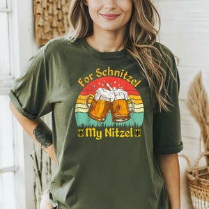 K&ouml;nnte beinhalten: Ein gr&uuml;nes T-Shirt mit einem Aufdruck von zwei Bierkr&uuml;gen, die ansto&szlig;en. Der Aufdruck ist vor einem Sonnenuntergangs-Hintergrund mit dem Text "For Schnitzel My Nitzel" platziert.