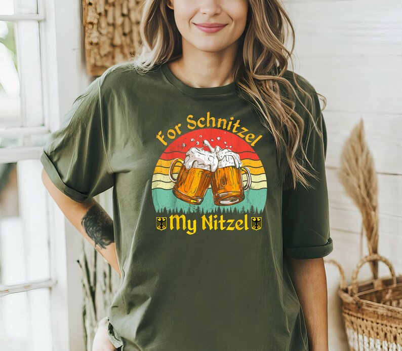 K&ouml;nnte beinhalten: Ein gr&uuml;nes T-Shirt mit einem Aufdruck von zwei Bierkr&uuml;gen, die ansto&szlig;en. Der Aufdruck ist vor einem Sonnenuntergangs-Hintergrund mit dem Text "For Schnitzel My Nitzel" platziert.