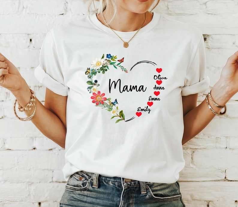May include: White t-shirt featuring a floral heart design with the word "Mama" in black script. The heart is adorned with colorful flowers, butterflies, and the names Olivia, Anna, Emma, and Emily.