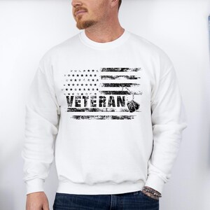 Peut inclure: Sweat-shirt blanc avec un motif de drapeau am&eacute;ricain vieilli et le mot "Veteran" imprim&eacute; en noir. Le motif du drapeau comprend une cha&icirc;ne avec des m&eacute;dailles.