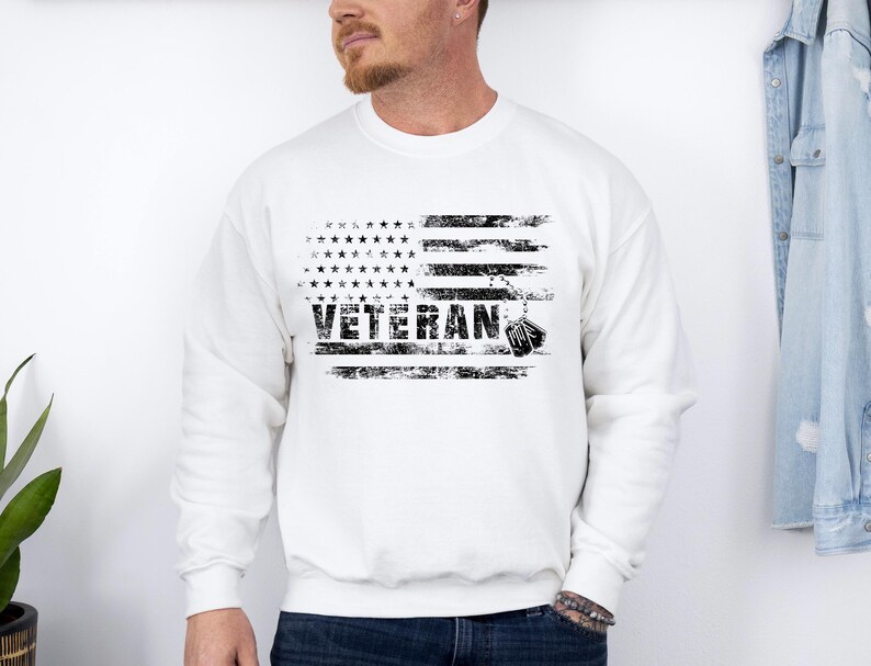 Peut inclure: Sweat-shirt blanc avec un motif de drapeau am&eacute;ricain vieilli et le mot "Veteran" imprim&eacute; en noir. Le motif du drapeau comprend une cha&icirc;ne avec des m&eacute;dailles.
