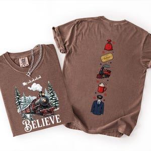 May include: A brown t-shirt featuring a graphic of a steam train in a snowy forest with the word "BELIEVE" below it. The back of the shirt displays a vertical arrangement of a red sack, a golden ticket, a train, a mug, and a conductor uniform.