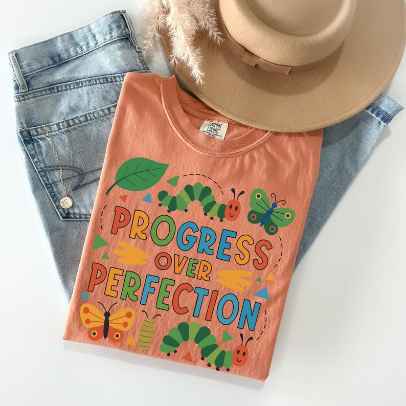 May include: An orange t-shirt with the words "PROGRESS OVER PERFECTION" in colorful letters, along with illustrations of caterpillars and butterflies. The shirt is paired with light blue jeans and a tan hat.