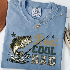 May include: A light blue t-shirt with a graphic of a fish jumping out of water, a fishing rod, and the words "Reel Cool Dad" in a camouflage font. A gold necklace is draped across the shirt.