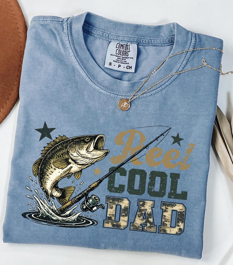 May include: A light blue t-shirt with a graphic of a fish jumping out of water, a fishing rod, and the words "Reel Cool Dad" in a camouflage font. A gold necklace is draped across the shirt.