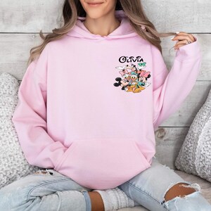 May include: Pink hoodie with a graphic of Mickey Mouse, Minnie Mouse, Donald Duck, Goofy, and Pluto. The text "Olivia" is printed on the hoodie.