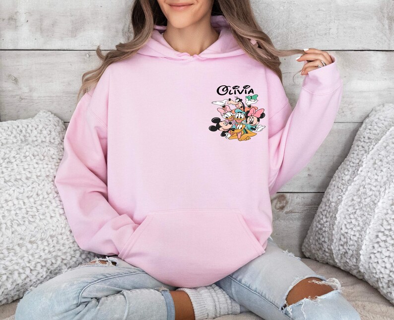 May include: Pink hoodie with a graphic of Mickey Mouse, Minnie Mouse, Donald Duck, Goofy, and Pluto. The text "Olivia" is printed on the hoodie.