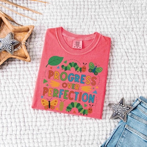 May include: A coral-colored t-shirt with the words "Progress Over Perfection" in a colorful, whimsical design featuring a leaf, caterpillars, and butterflies. The shirt is laid flat on a textured white surface.
