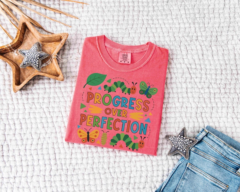 May include: A coral-colored t-shirt with the words "Progress Over Perfection" in a colorful, whimsical design featuring a leaf, caterpillars, and butterflies. The shirt is laid flat on a textured white surface.