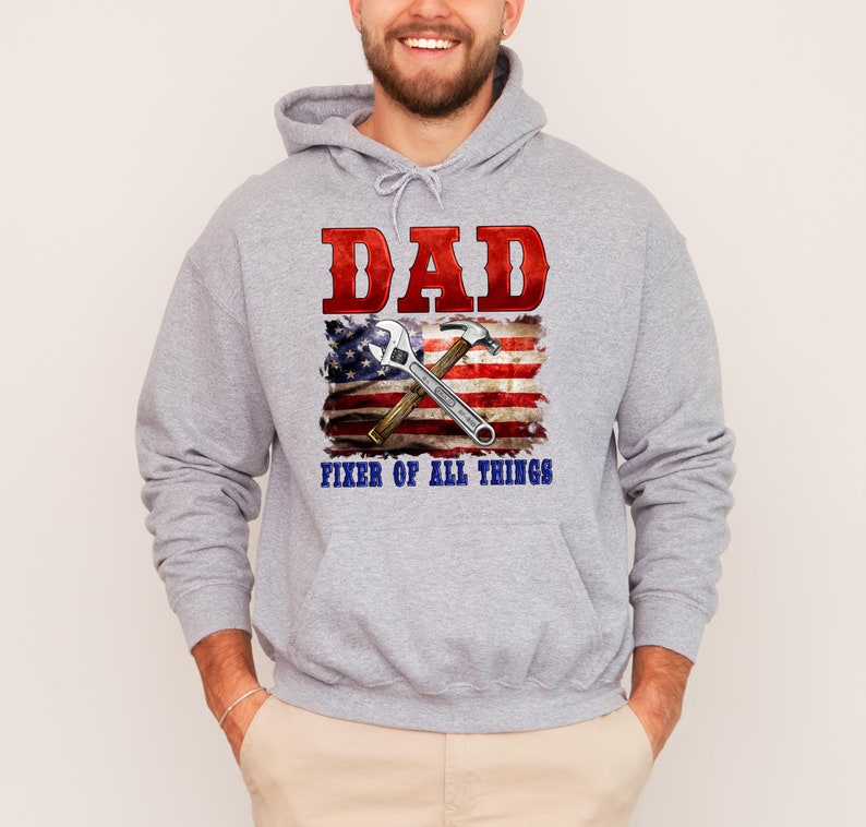 May include: Gray hooded sweatshirt with the text "DAD FIXER OF ALL THINGS" in red and blue. An American flag with a wrench and hammer is behind the text.