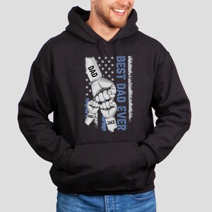 May include: Black hooded sweatshirt with a graphic of three fists raised in the air, with the words "Best Dad Ever" and the names "Dad", "Sander", and "Jack" printed on the fists. The graphic is set against a blue and white American flag background.