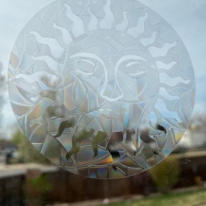 May include: A clear glass sun with a face and closed eyes. The sun is etched with a geometric pattern and has a rainbow iridescent effect.
