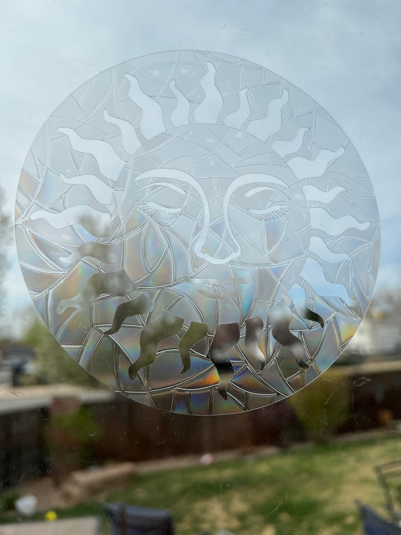 May include: A clear glass sun with a face and closed eyes. The sun is etched with a geometric pattern and has a rainbow iridescent effect.