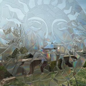 May include: A clear glass window with a decorative sun design etched into the surface. The sun has a face with closed eyes and a geometric pattern around it. The design is iridescent and reflects light.
