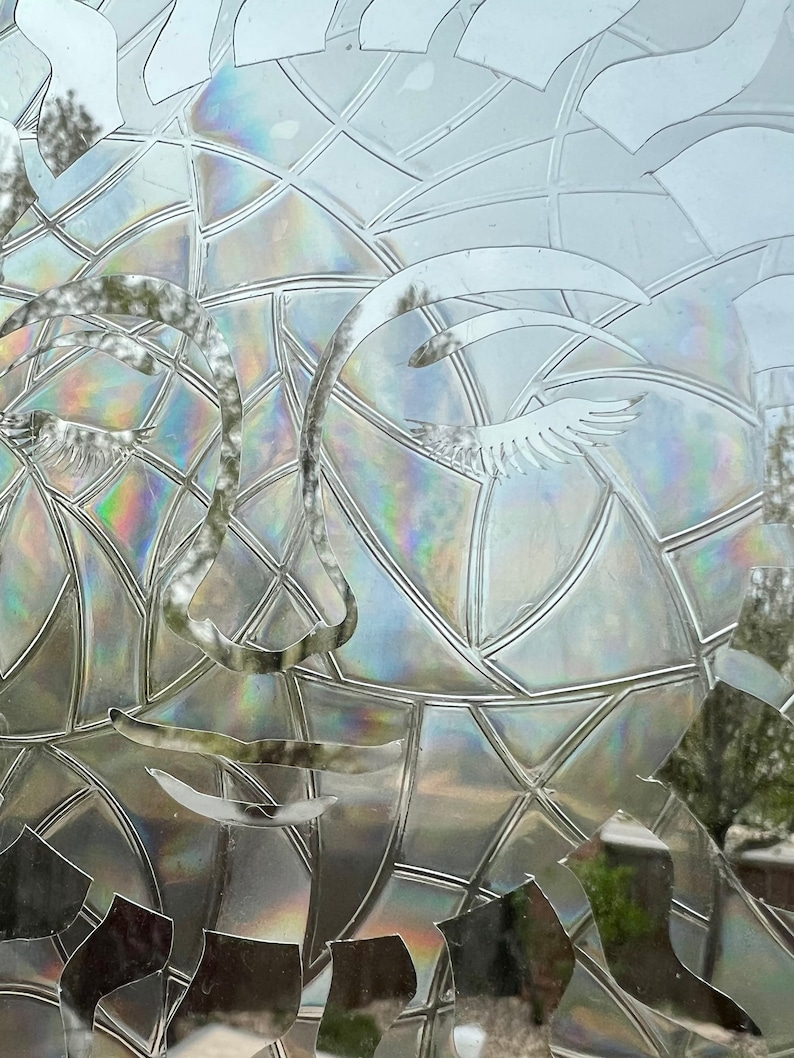 May include: A close-up of a clear glass window with a decorative film applied. The film features a geometric pattern that creates the illusion of a face with eyelashes and a nose.