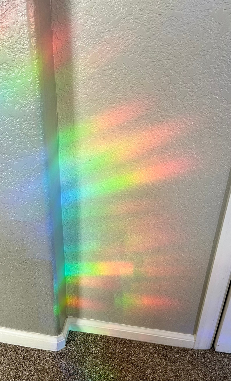 May include: A rainbow prism hanging in a window casts colorful light onto a white wall with textured paint. The light creates a pattern of overlapping bands of red, orange, yellow, green, blue, indigo, and violet.