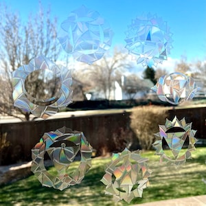 May include: Seven iridescent chakra symbols, each with a unique design, are displayed on a window. The symbols are made of a reflective material that creates a rainbow effect.