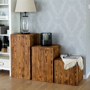 Puede incluir: Tres taburetes de madera en forma de cubo de diferentes tama&ntilde;os, apilados en forma de escalera. Los taburetes son de color marr&oacute;n oscuro con un patr&oacute;n de veta de madera r&uacute;stico. El taburete m&aacute;s grande est&aacute; a la izquierda, el m&aacute;s peque&ntilde;o a la derecha.