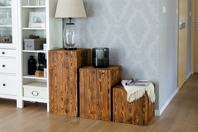 Puede incluir: Tres taburetes de madera en forma de cubo de diferentes tama&ntilde;os, apilados en forma de escalera. Los taburetes son de color marr&oacute;n oscuro con un patr&oacute;n de veta de madera r&uacute;stico. El taburete m&aacute;s grande est&aacute; a la izquierda, el m&aacute;s peque&ntilde;o a la derecha.