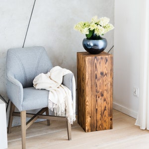 Puede incluir: Un pedestal de madera con un acabado marr&oacute;n oscuro y un aspecto desgastado. Un jarr&oacute;n con flores blancas se coloca encima del pedestal. Un sill&oacute;n gris con una manta blanca se encuentra en el fondo.