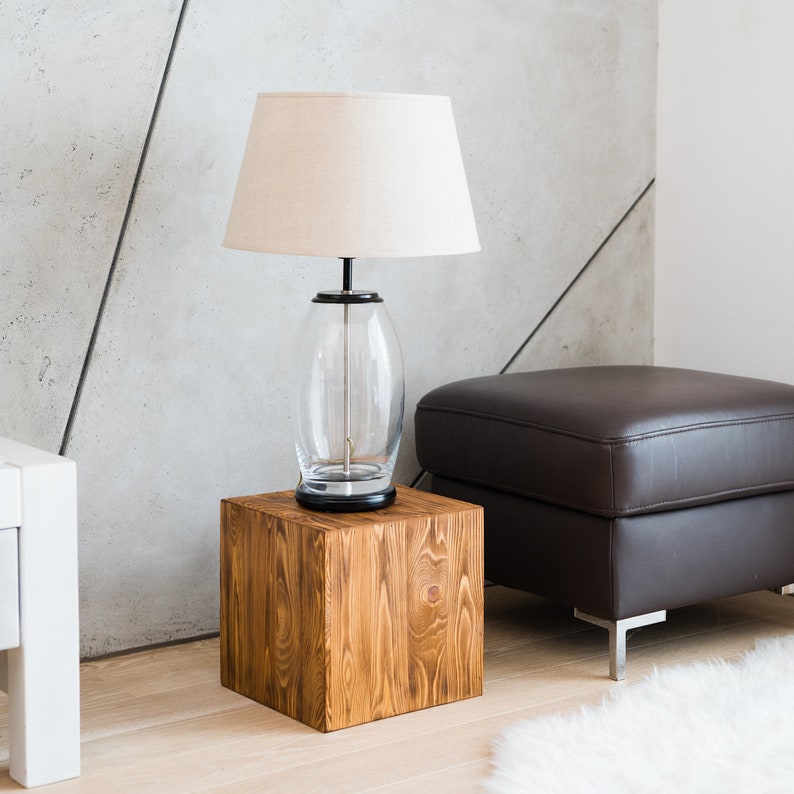 Puede incluir: Una pantalla de l&aacute;mpara blanca con una base de vidrio transparente se asienta sobre una mesa auxiliar de madera. La mesa est&aacute; junto a un otomano de cuero marr&oacute;n.