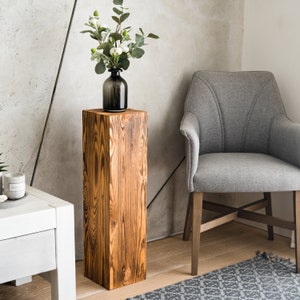 Puede incluir: Un pedestal de madera r&uacute;stico con un acabado de madera natural. El pedestal es alto y rectangular, con una superficie lisa. Un jarr&oacute;n con flores blancas se coloca encima del pedestal.