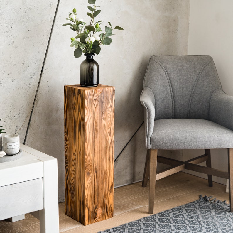 Puede incluir: Un pedestal de madera r&uacute;stico con un acabado de madera natural. El pedestal es alto y rectangular, con una superficie lisa. Un jarr&oacute;n con flores blancas se coloca encima del pedestal.