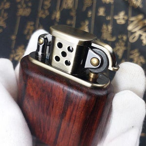 May include: A wooden lighter with a brass top and a metal flint wheel. The lighter has a dark wood grain pattern and a polished brass finish.