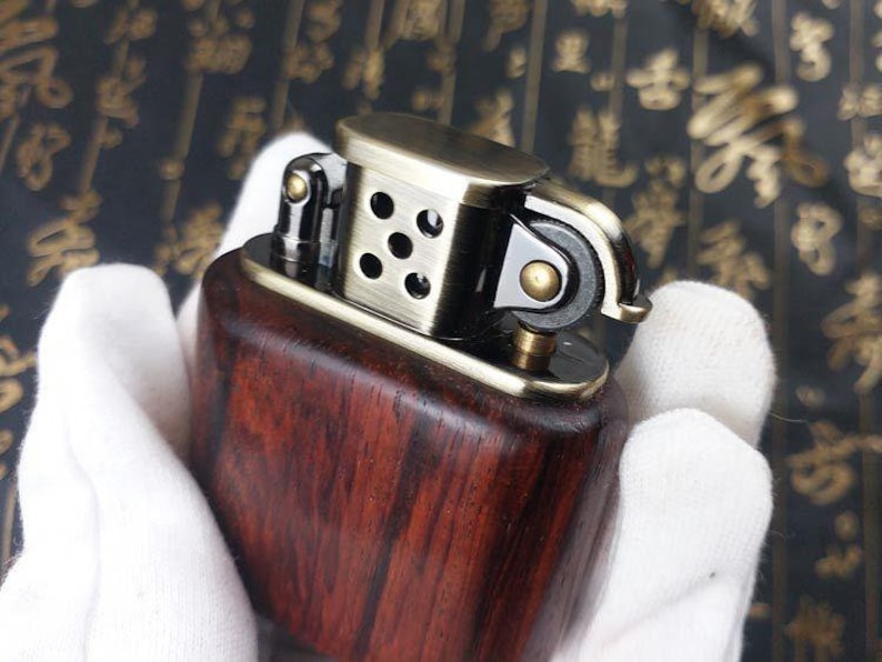 May include: A wooden lighter with a brass top and a metal flint wheel. The lighter has a dark wood grain pattern and a polished brass finish.