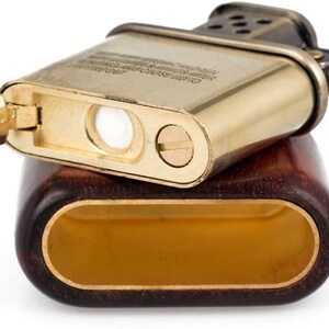 May include: A gold-toned lighter with a wooden case. The lighter has a wick and a flint, and the case has a dark brown wood grain.