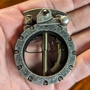 May include: A silver-toned lighter with a round, clear window revealing a wick and a metal rod. The lighter has a textured surface and a lever on the side.