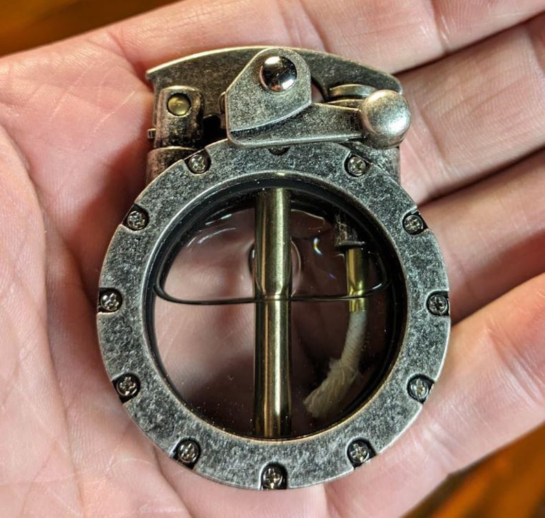 May include: A silver-toned lighter with a round, clear window revealing a wick and a metal rod. The lighter has a textured surface and a lever on the side.