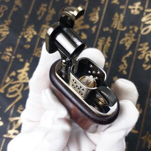 May include: A wooden and metal lighter with a silver and black finish. The lighter has a small, round, silver wick and a black, rectangular body.