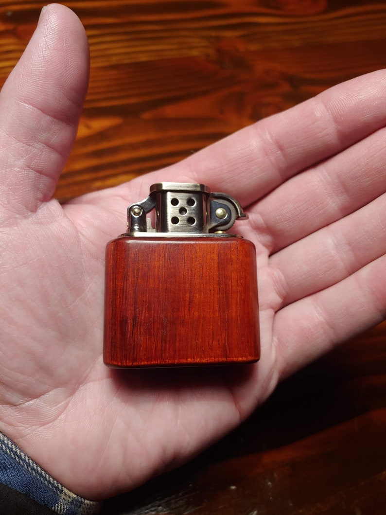 May include: A wooden lighter with a silver top and a red wood finish. The lighter is in the palm of a hand.