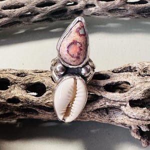May include: A silver ring featuring a teardrop-shaped stone with pink and orange hues set above a white cowrie shell. The metal setting includes small silver spheres between the stone and the shell, creating a layered, vertical design.