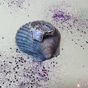 Peut inclure: Une bague en argent ornée d'une pierre précieuse en forme d'étoile, de couleur bleu clair, est posée sur un coquillage bleu. La bague est posée sur un fond vert clair parsemé de paillettes violettes. La bande de la bague a un motif décoratif.