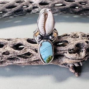 May include: A silver ring featuring a vertically oriented white cowrie shell at the top and a smooth, oval-shaped blue and green stone at the bottom. The setting includes small silver spheres between the shell and the stone, with a polished metal band.