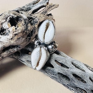 May include: A silver ring featuring two white cowrie shells set vertically. The metal band has a polished finish with small silver spheres positioned between the shells. The design showcases a dual-shell arrangement secured within a silver setting.