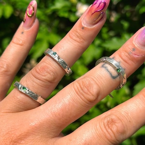 May include: Three silver rings with textured bands and small, green gemstones. The rings are worn on fingers with colourful nail art. The background is a blurred view of green foliage.