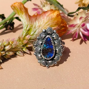 May include: A silver ring with a teardrop-shaped opal stone. The opal has a vibrant blue and green color. The ring is surrounded by a detailed, ornate setting with a floral design. The background includes dried flowers.