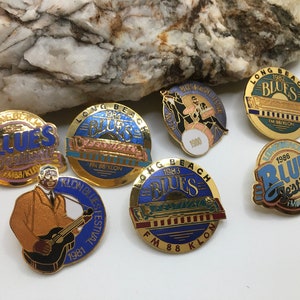 Puede incluir: Una colección de siete pines de color dorado con el Festival de Blues de Long Beach. Los pines son circulares y presentan diferentes años, incluyendo 1980, 1981, 1982, 1983, 1984, 1986. Un pin presenta una imagen de dibujos animados de un músico tocando la guitarra.
