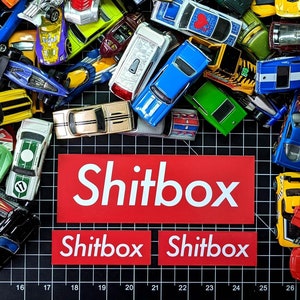 May include: A collection of toy cars in various colors and styles, including red, blue, green, yellow, and black. The cars are arranged in a random pattern on a black and white grid background. The words "Shitbox" are printed in white letters on a red background.
