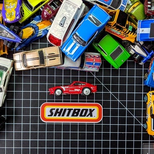 Pode incluir: Uma coleção de carrinhos de brinquedo em várias cores, incluindo vermelho, azul, verde e amarelo, dispostos em um fundo de grade preta. Os carrinhos são de diferentes marcas e modelos, e alguns têm detalhes únicos como chamas ou números de corrida. O texto "SHITBOX" está impresso em um adesivo vermelho e branco.