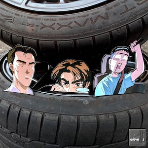 Può includere: Primo piano di uno pneumatico con tre personaggi dei cartoni animati dell'anime Initial D. I personaggi sono Takumi Fujiwara, Itsuki Takeuchi e Keisuke Takahashi. Lo pneumatico ha il testo "MAXXI" e "10021105" stampato su di esso.